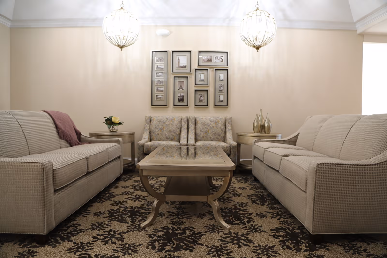 A cozy living room with two beige sofas facing each other, a patterned armchair in the center back, a wooden coffee table in the middle, two round side tables with decorative items, a patterned carpet, and two hanging pendant lights. A wall with framed pictures is visible behind the armchair.