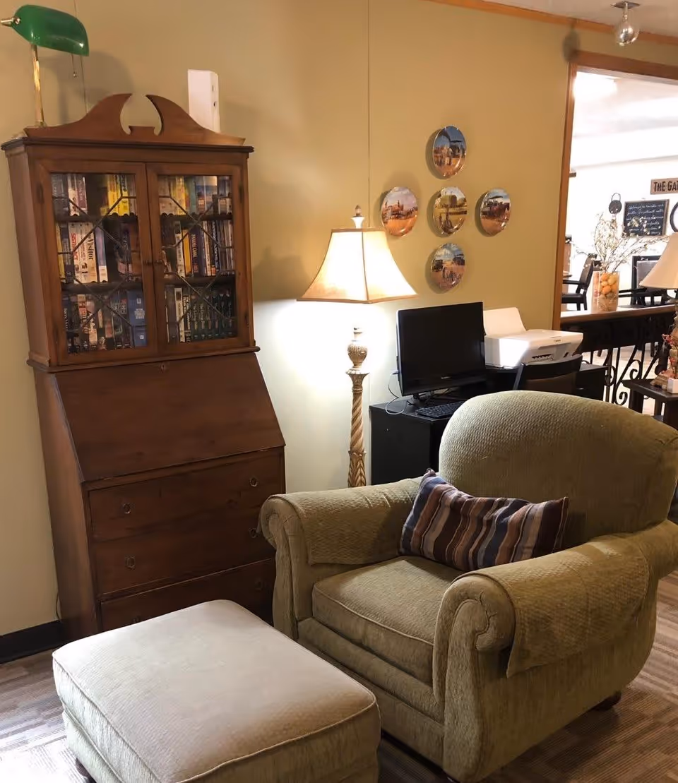 Cozy interior corner of a retirement home featuring a green upholstered armchair with a striped pillow, a matching ottoman, a wooden cabinet with glass doors filled with books, a tall floor lamp with a beige shade, and a small desk area with a computer monitor and printer. Decorative plates are mounted on the wall, and a glimpse of a dining area is visible in the background.