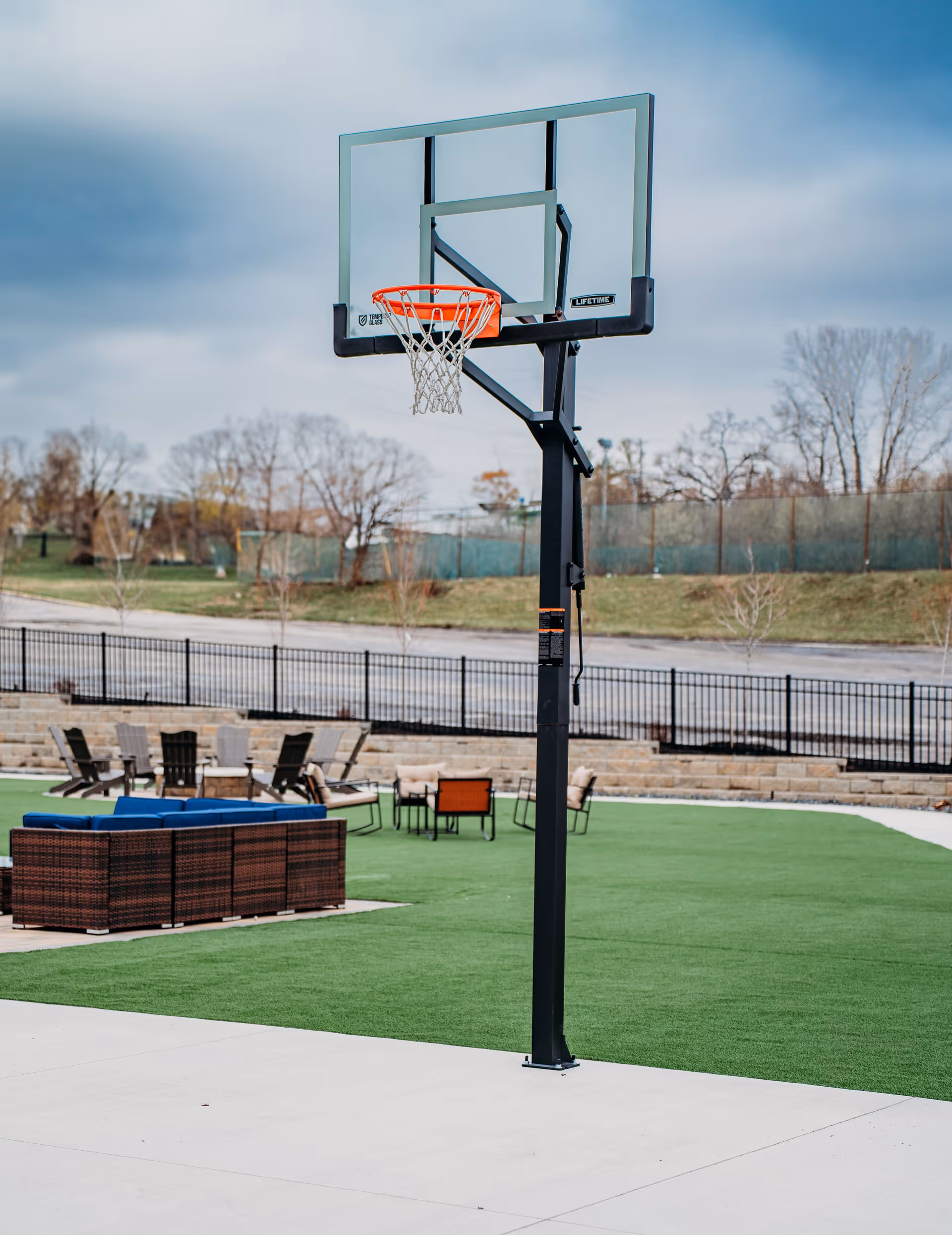 Outdoor basketball hoop on a concrete court with green artificial turf and patio furniture including chairs and sofas in the background, surrounded by a black metal fence and leafless trees under a cloudy sky.