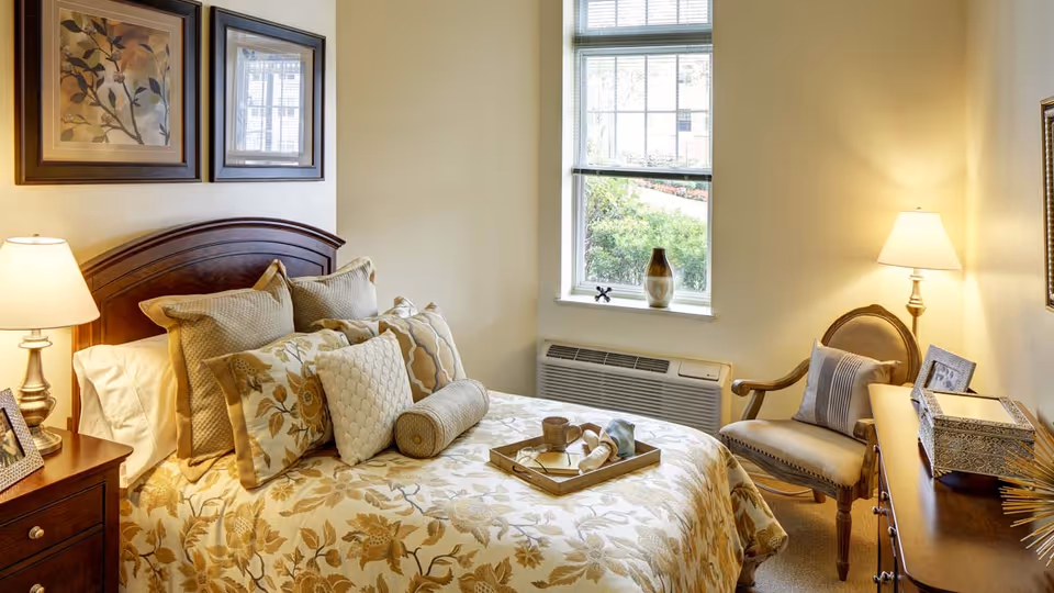 A cozy bedroom with a wooden headboard bed adorned with multiple decorative pillows and a floral bedspread. There is a wooden nightstand with a lamp and a framed photo on the left side of the bed. A window with blinds lets in natural light, and a wooden chair with a cushion is placed next to a wooden dresser with a lamp and decorative items.