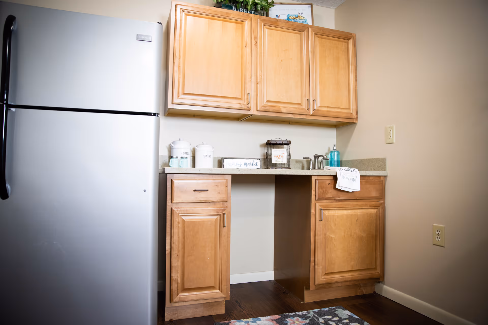 A small kitchen area with light wooden cabinets, a stainless steel refrigerator on the left, a countertop with a sink, soap dispenser, and decorative containers. There is a floral patterned rug on the dark wooden floor.