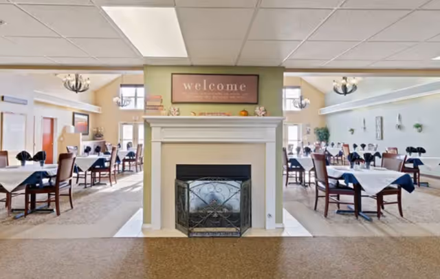 Spacious senior dining room with a central decorative fireplace and multiple tables set with white tablecloths and dark napkins.