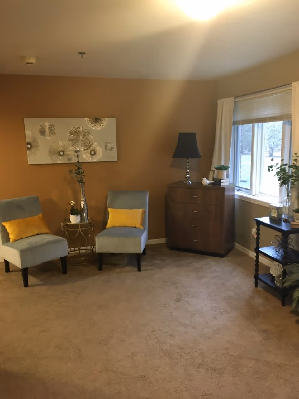 Cozy living room with two gray accent chairs with yellow pillows, side tables, a wooden dresser with a lamp, wall art, and a window.