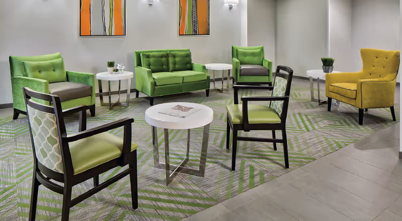 A modern seating area with green and yellow armchairs and a green loveseat arranged around white round tables on a patterned carpet. Two abstract orange and green paintings hang on the wall behind the seating.