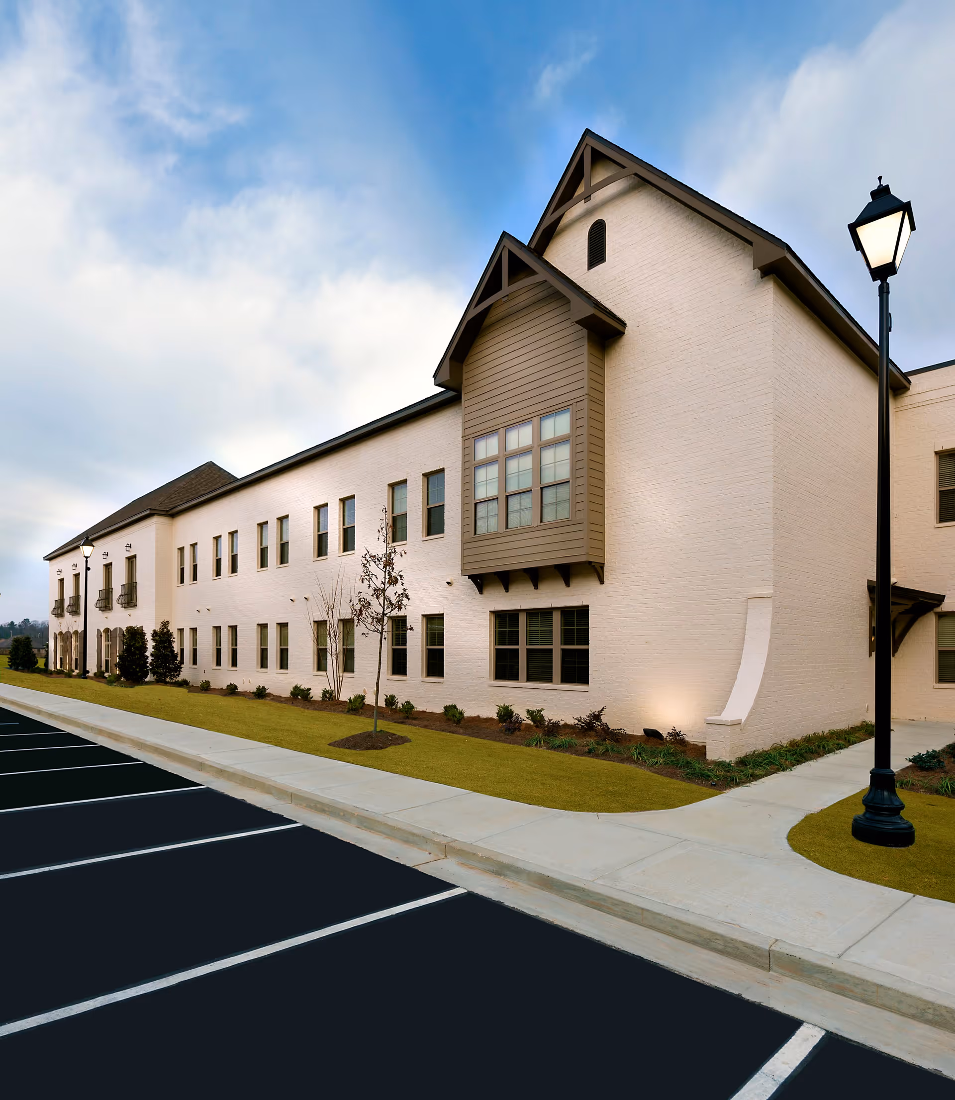 Exterior view of a two-story senior living facility building with beige brick walls, multiple windows, small landscaped areas with shrubs and young trees, a sidewalk, and a black street lamp. The sky is partly cloudy.