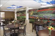 Interior view of a dining area with several round tables and chairs. Each table has a small vase with flowers. The walls feature a large mural depicting a colorful garden with rows of flowers and greenery. The ceiling has panels with sky and cloud images, adding a bright and airy feel to the room.