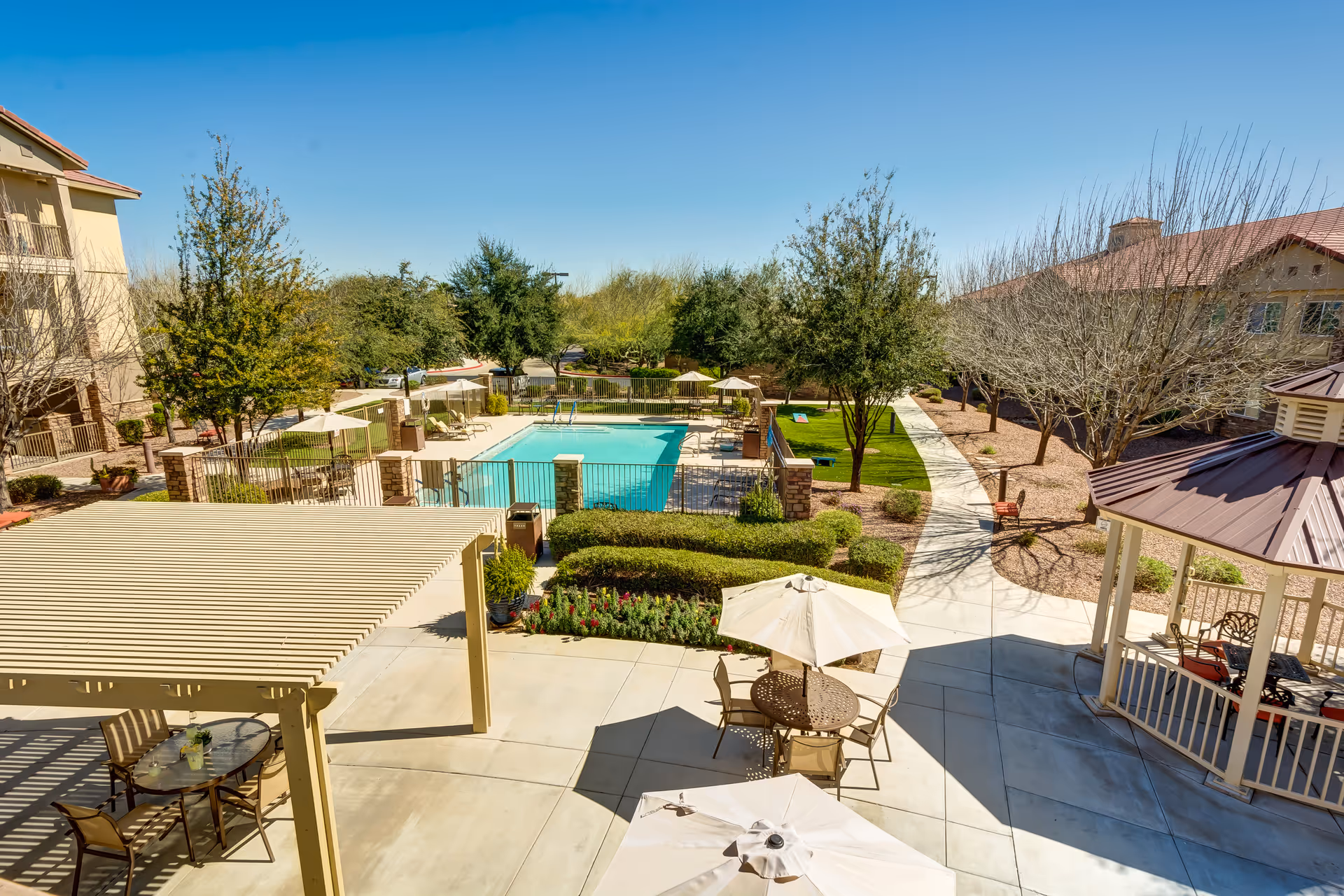 Outdoor area of a senior living facility featuring a swimming pool surrounded by lounge chairs and umbrellas, a gazebo with seating, a pergola with a table and chairs, landscaped greenery, and a paved walkway under a clear blue sky.