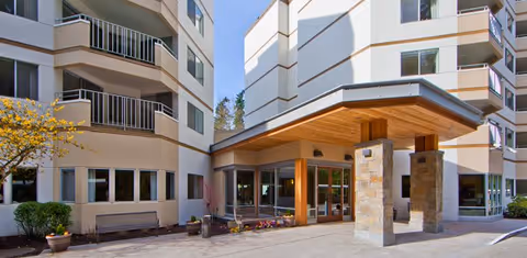 Exterior view of a multi-story senior living facility with balconies, large windows, and a covered entrance supported by stone pillars. There are potted plants and a small tree near the entrance under a clear blue sky.