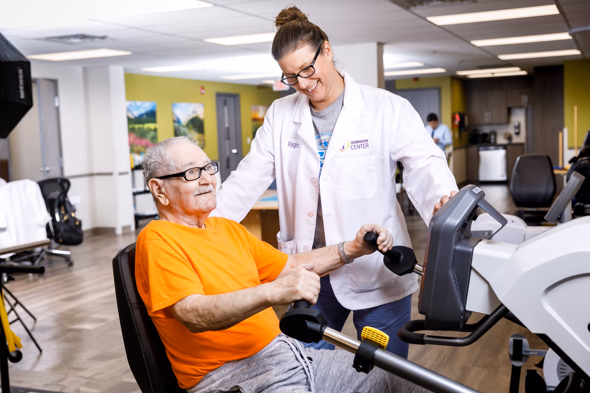 An elderly man wearing an orange shirt is using a seated exercise machine in a rehabilitation or therapy room. A female healthcare professional in a white coat labeled Hammonton Center is standing beside him, smiling and providing support. The room has wooden flooring, exercise equipment, and colorful wall art in the background.