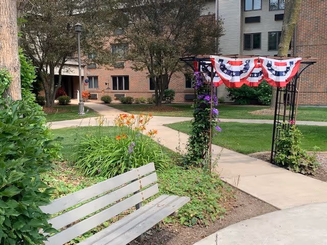 Courtyard with a bench, flower beds, paved walkways and a pergola draped in patriotic bunting in front of a multi-story brick building.