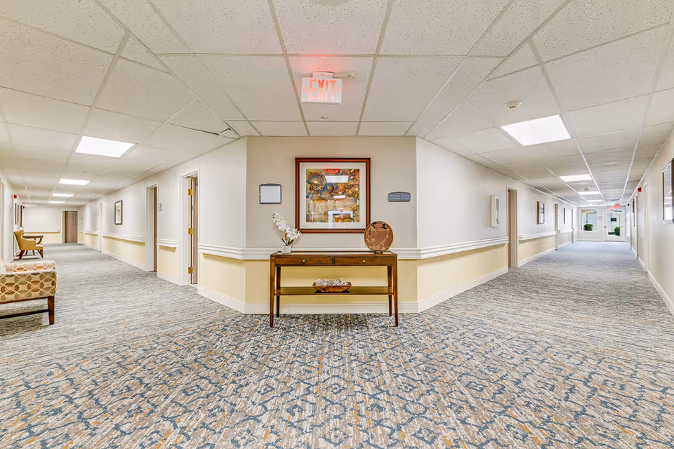 Carpeted interior hallway in a senior living facility with a console table and framed art at the center, seating and doors along both sides, and an exit sign overhead.