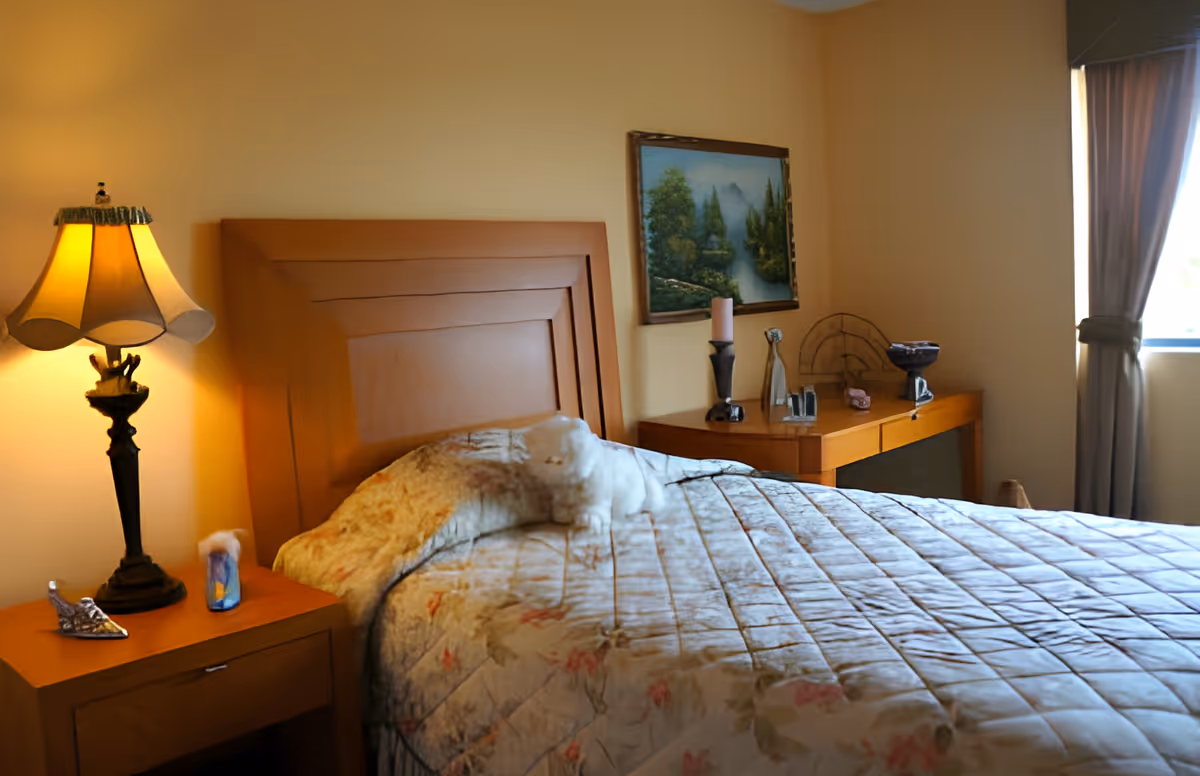 A cozy bedroom with a wooden headboard and a floral quilted bedspread. A bedside table with a decorative lamp and small items is on the left. On the right, there is a wooden dresser with various decorative objects and a framed landscape painting hanging above it. A window with curtains allows natural light into the room.