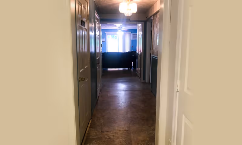 View down a narrow hallway with doors on both sides opening to a living area with a couch and windows at the far end.