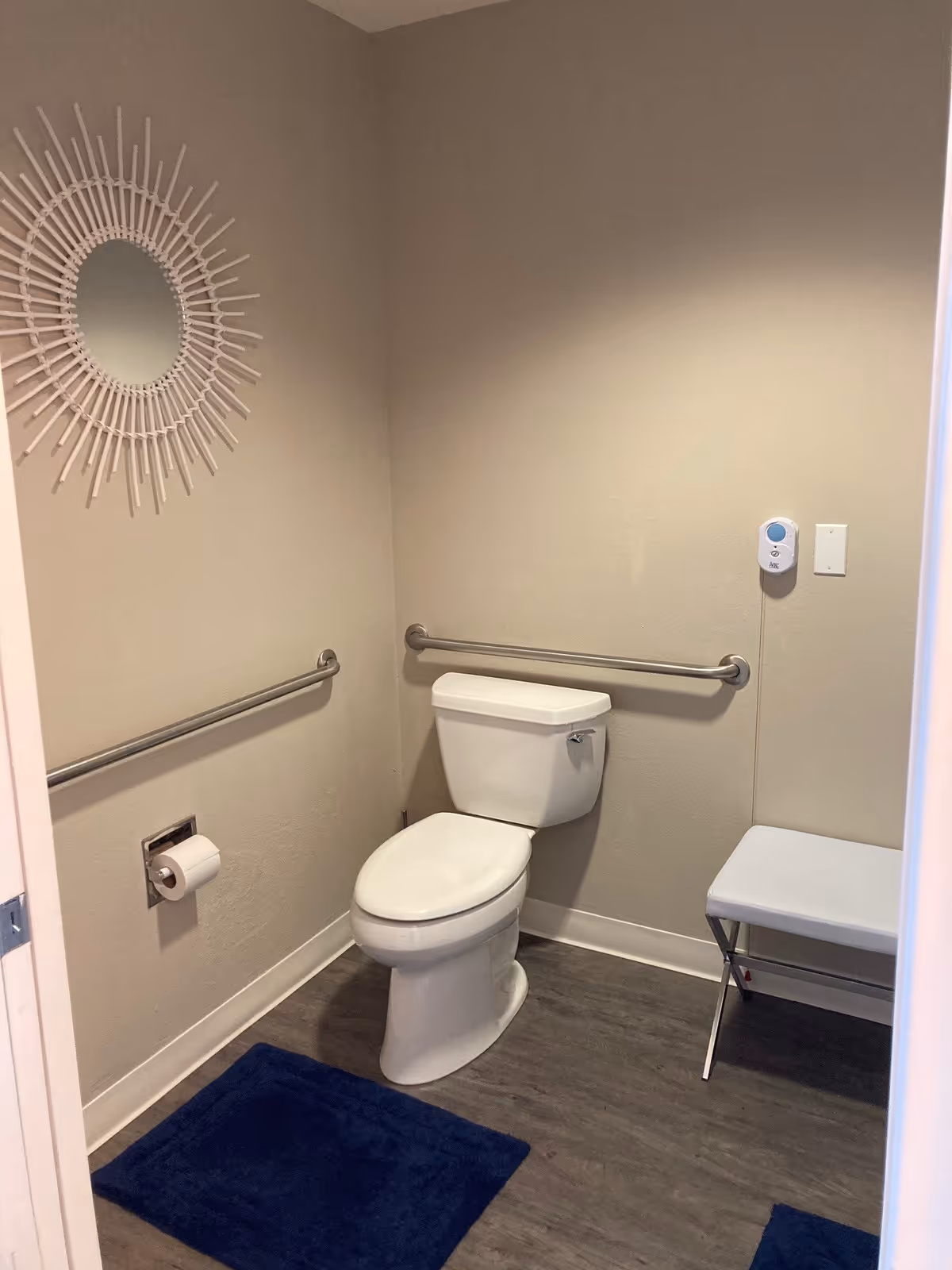 Small accessible bathroom with a toilet, horizontal grab bars, blue rugs, a decorative round mirror, and a small folding bench.