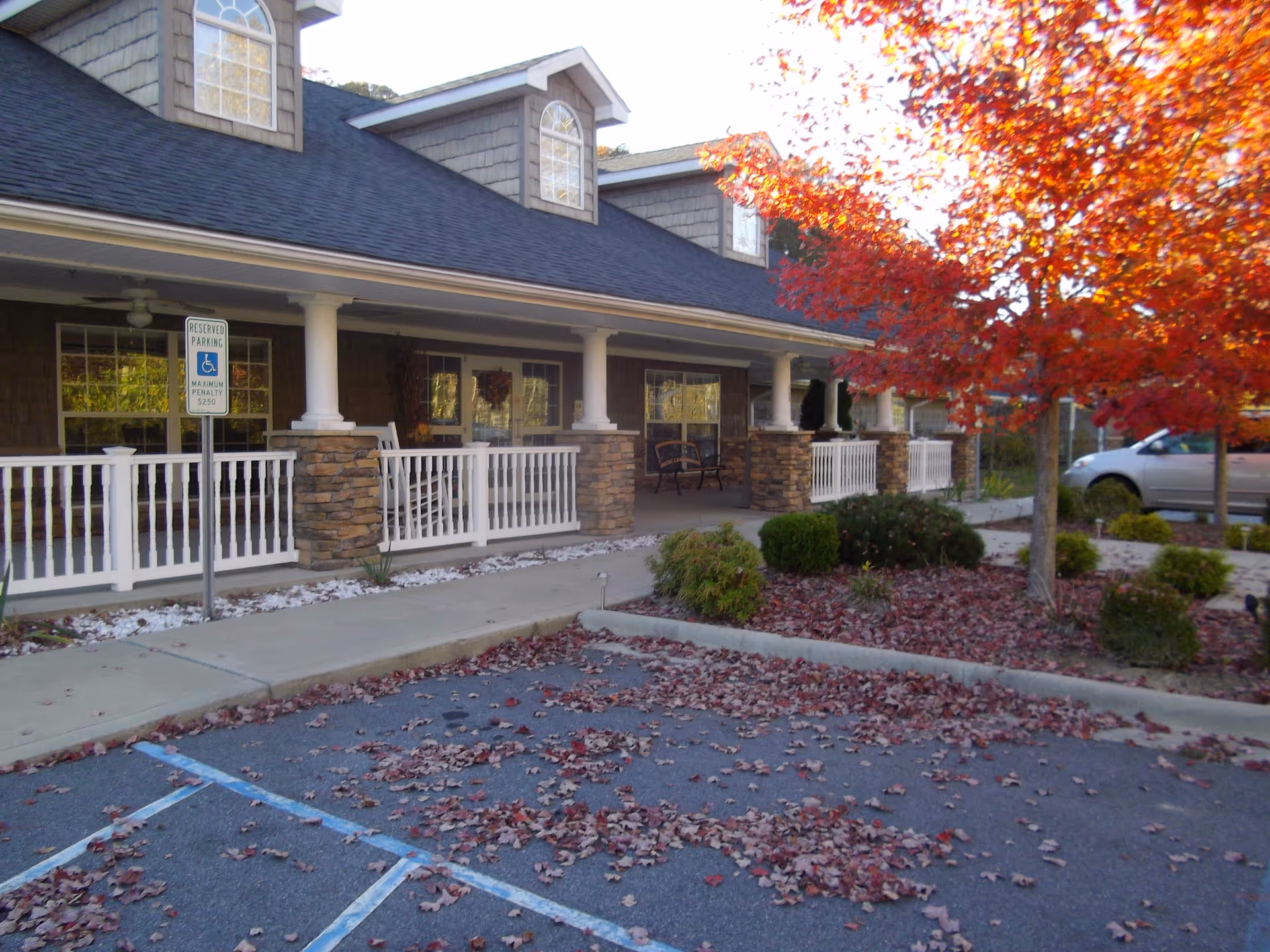 Front exterior of a single-story building with a covered porch, white railing and columns, a handicap parking sign, and a red-leaved tree in the foreground.