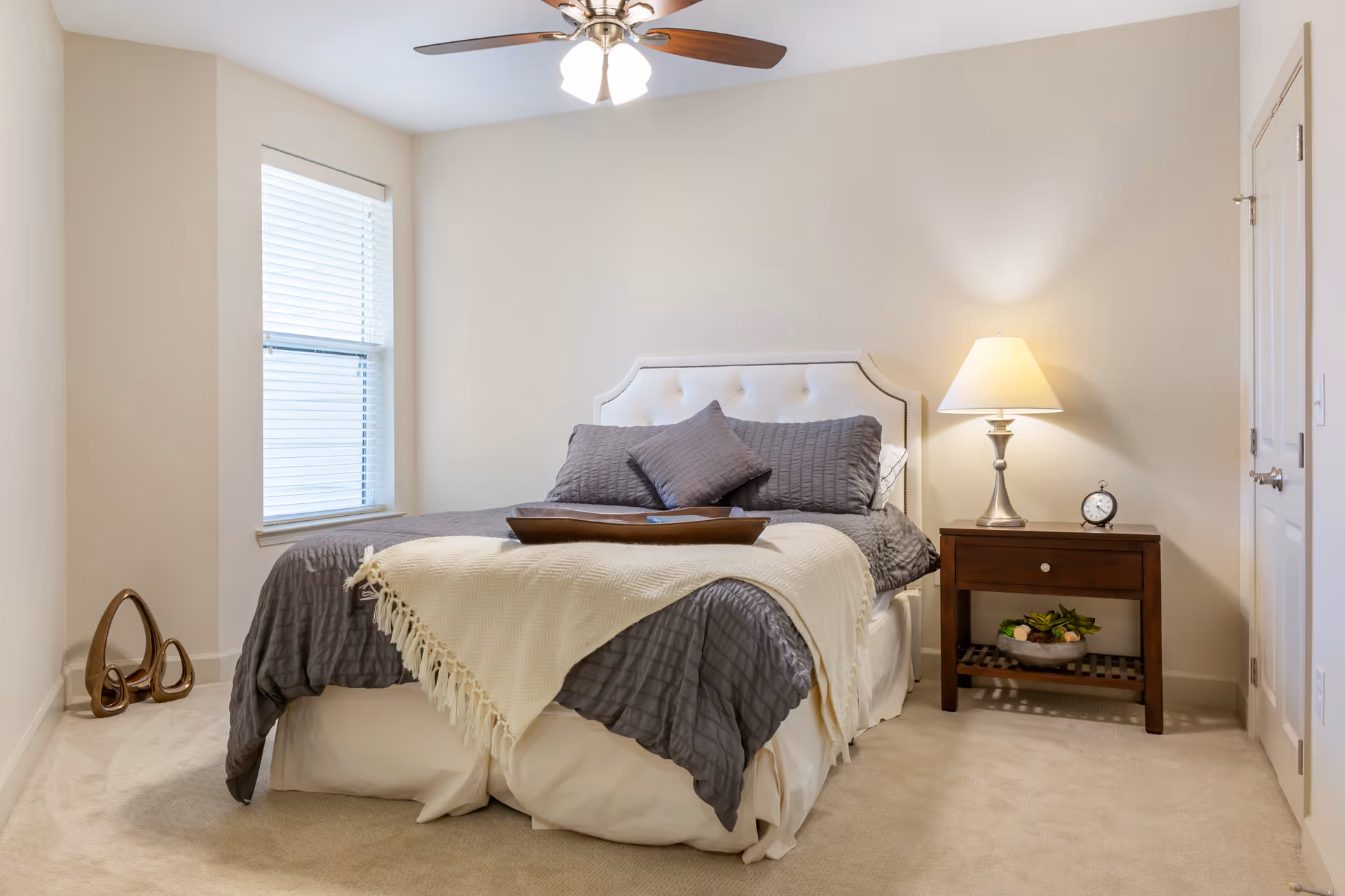 A neatly made bedroom with a bed, pillows, nightstand with lamp and clock, window, and ceiling fan.