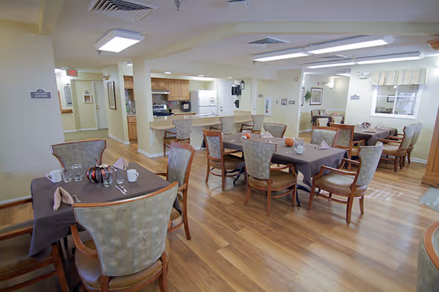 Well-lit dining room with several set tables and chairs and a view into a kitchen area.