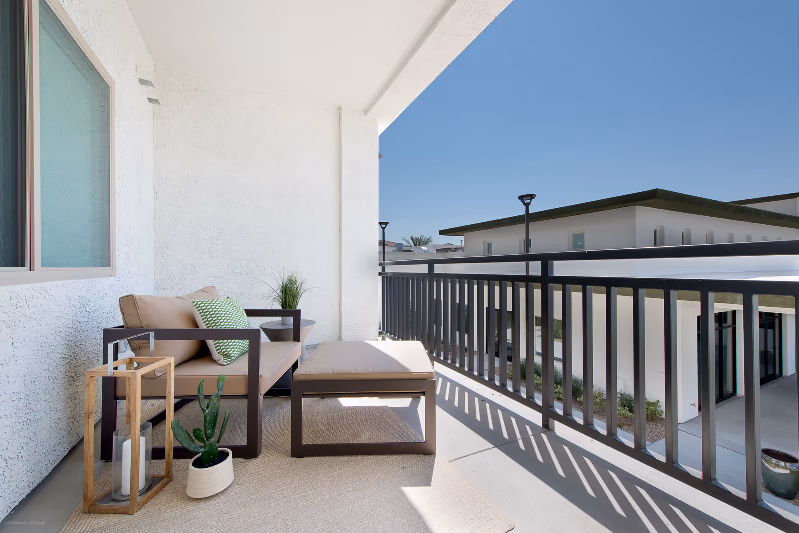 Sunlit balcony with cushioned outdoor seating, ottoman, small plants, and a railing overlooking neighboring buildings.