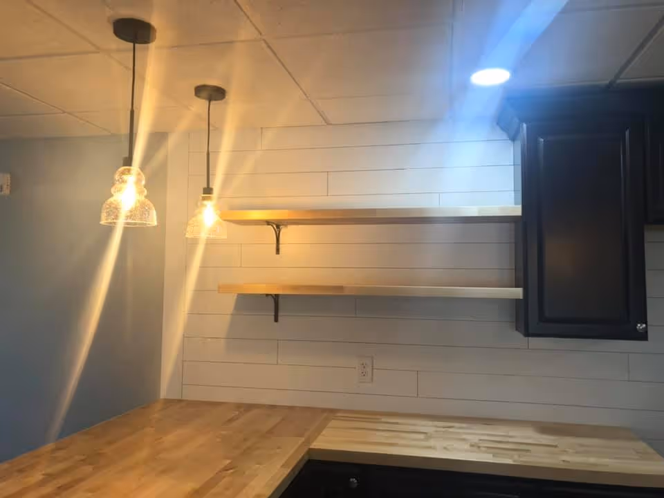 Kitchen corner with wooden countertops, two floating wood shelves on a shiplap wall, a dark cabinet, and pendant lights.