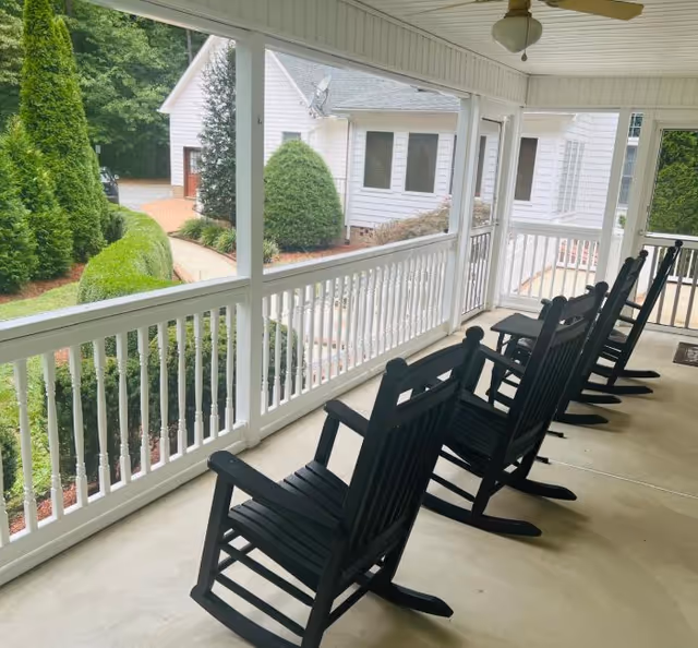 Covered porch with a row of black rocking chairs overlooking landscaped grounds and an adjacent building.