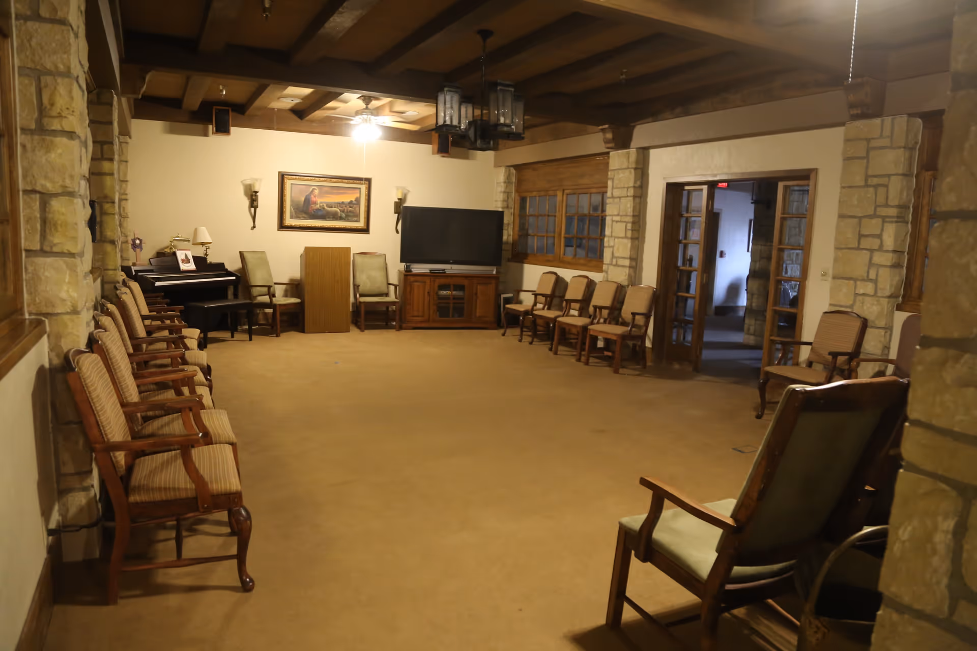 Large communal sitting room with rows of wooden chairs lining a carpeted space, a piano, TV, and wood-beamed ceiling.