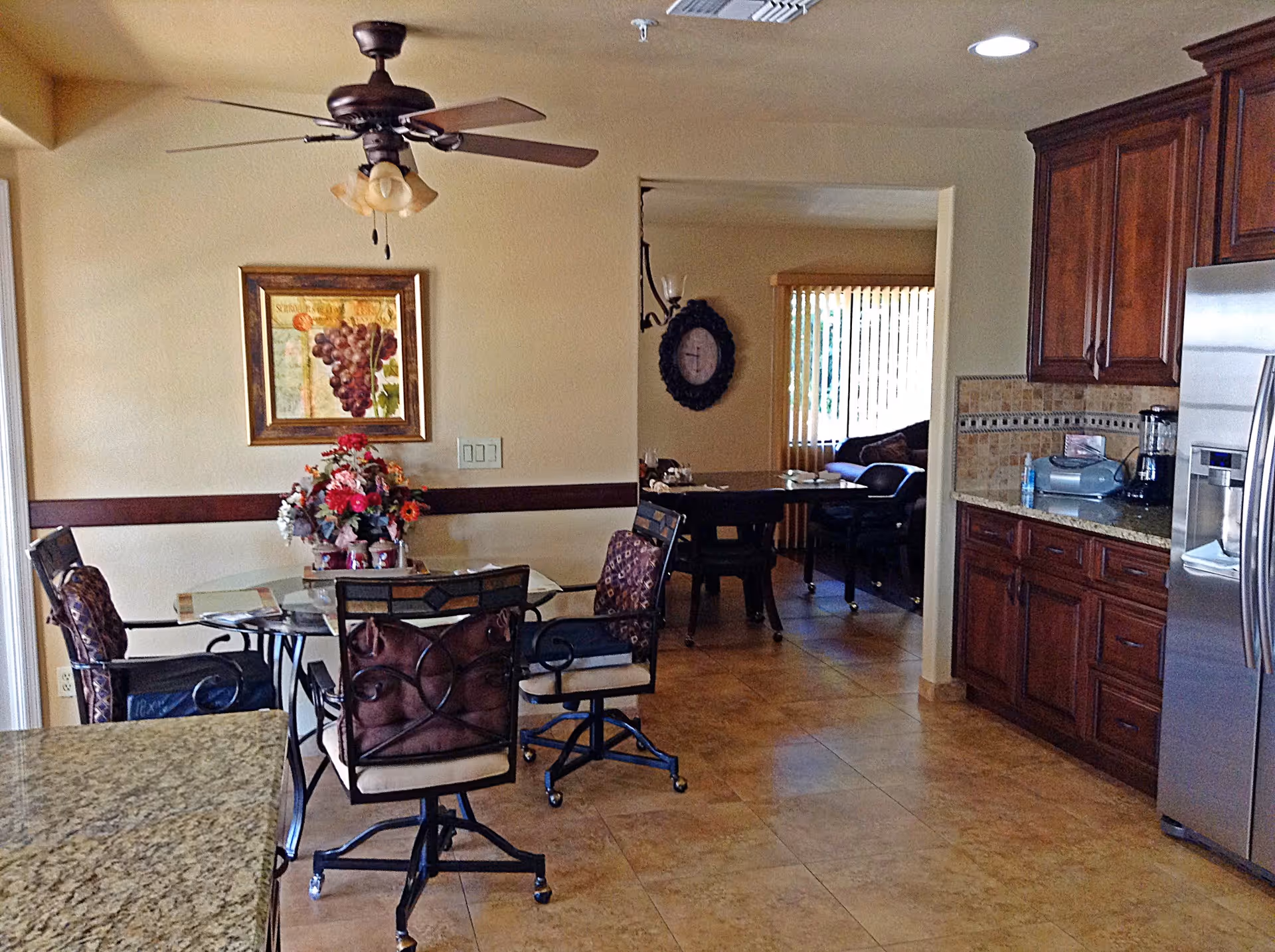 Interior view of a dining area with a glass-top table surrounded by four cushioned chairs on wheels. A ceiling fan with lights hangs above the table. There is a framed picture of grapes on the wall and a floral centerpiece on the table. To the right, there is a kitchen area with wooden cabinets, a granite countertop, and stainless steel appliances. In the background, another room with a large clock on the wall and a window with vertical blinds is visible.