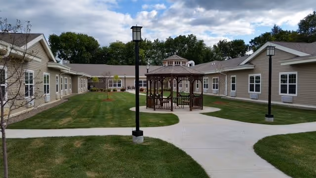 Outdoor courtyard area of Homestead Assisted Living & Memory Care of Morrison featuring a central wooden gazebo with benches, surrounded by a paved walkway, green lawn, and single-story beige buildings with white-trimmed windows under a partly cloudy sky.