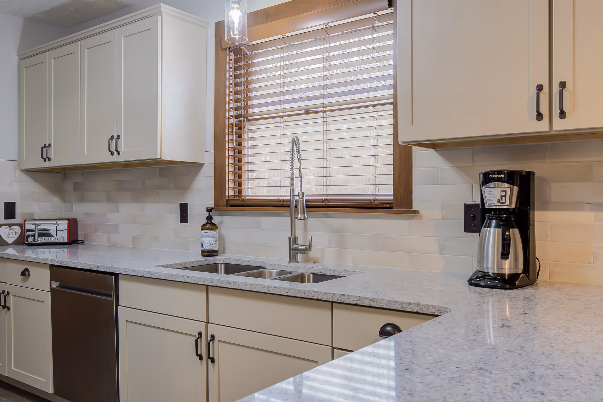 Modern kitchen interior with white cabinets, a double sink under a window with wooden blinds, a coffee maker on the right countertop, a toaster on the left countertop, and a soap dispenser near the sink.