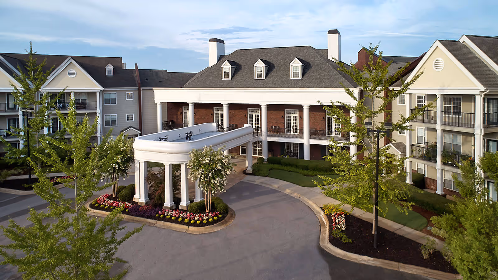 Front exterior view of Fairview At Redstone Village, showing a large building with multiple floors, white columns, balconies, and a covered driveway entrance surrounded by landscaped greenery and flower beds.