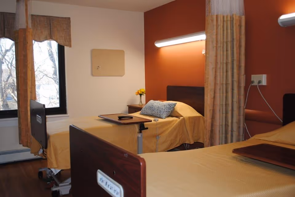 A room with two hospital-style beds separated by a privacy curtain. Each bed has a yellow bedspread and a wooden tray table. One bed has a blue patterned pillow and a small vase with yellow flowers on a bedside table. There is a window with brown curtains on the left side and a wall-mounted light above one bed.
