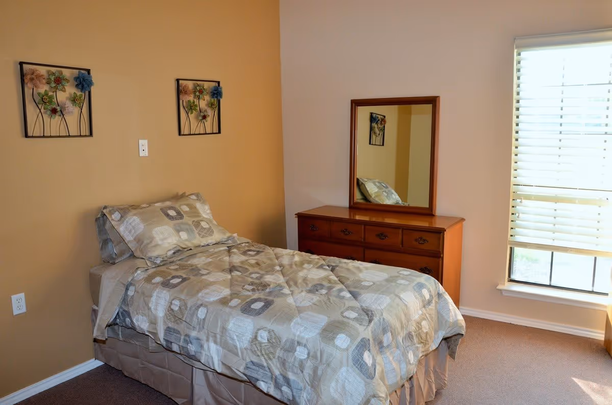 A bedroom with a single bed covered in a beige and gray patterned comforter and pillow. The room has a two-tone wall with one side painted light brown and the other white. Above the bed are two decorative floral wall hangings. Next to the bed is a wooden dresser with a mirror reflecting part of the bed and wall decor. A window with white blinds lets in natural light.
