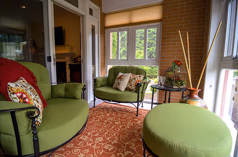 Sunroom sitting area with green cushioned chairs and ottoman, a patterned rug, side table with decor, and large windows.