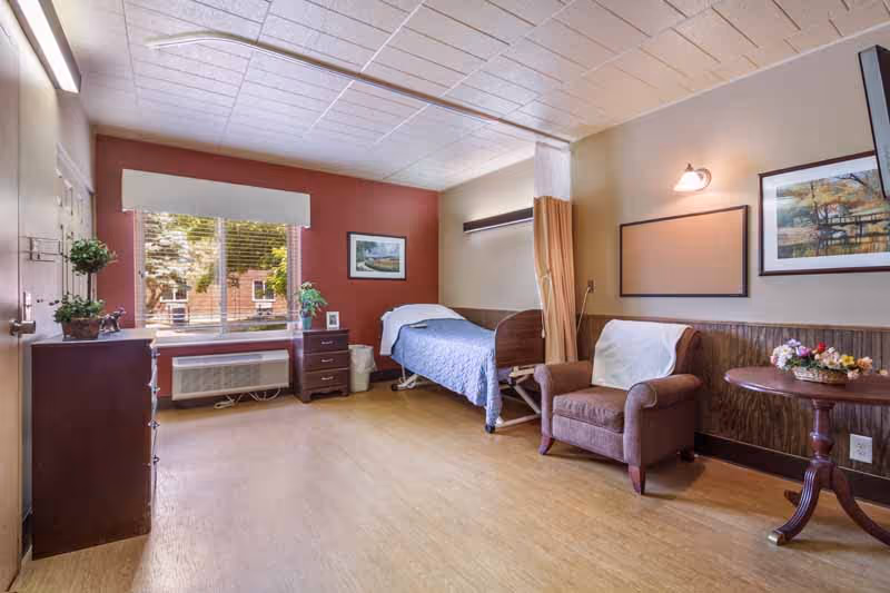 Well-lit nursing facility patient room with a single bed, recliner chair, dresser, and a window overlooking trees.
