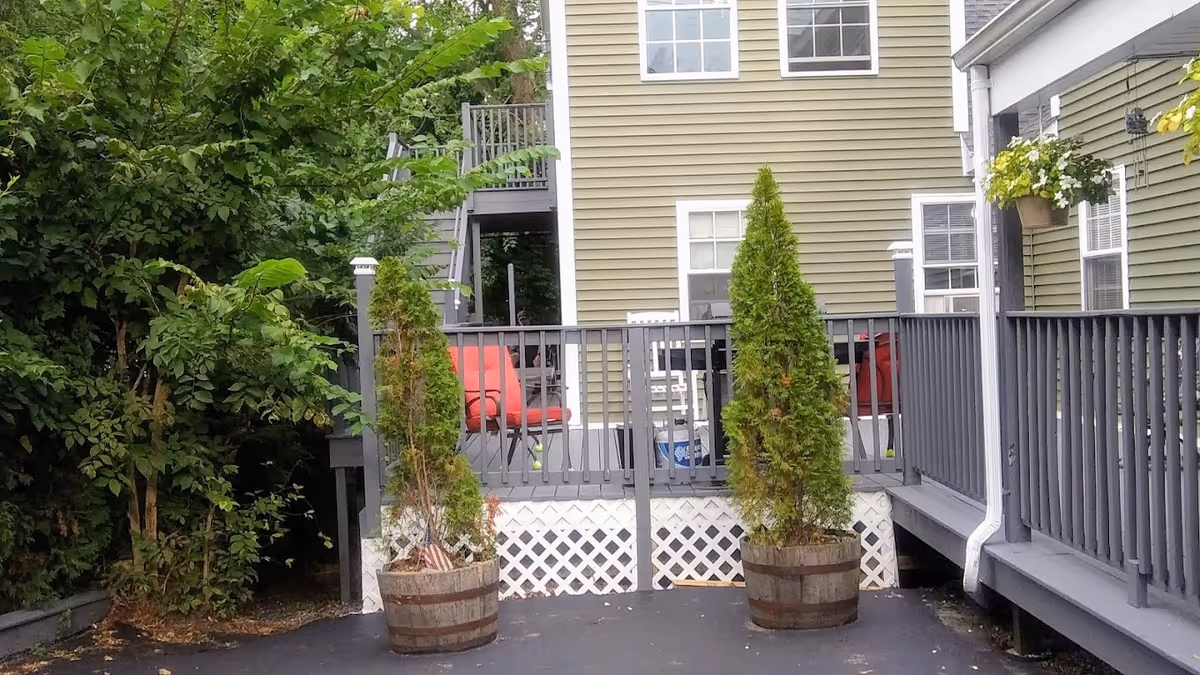 Wooden deck and railing attached to a green-sided building with two potted evergreen trees and red patio chairs.