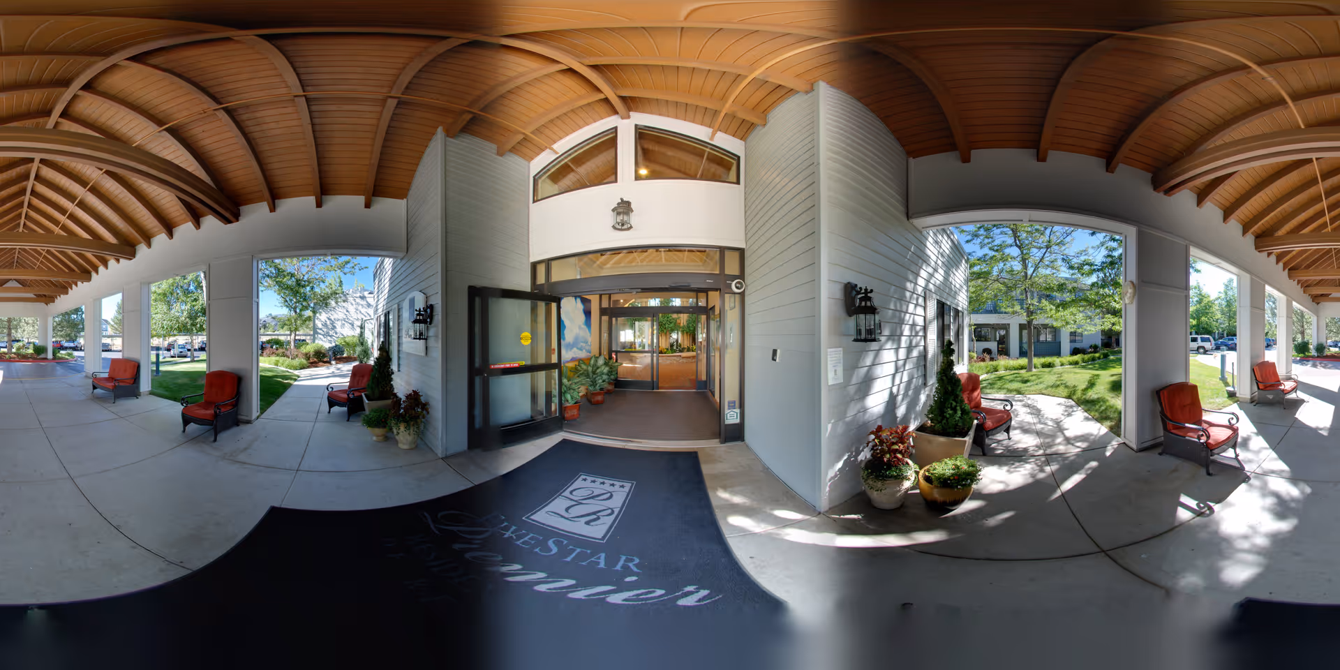 Covered entrance area of Five Star Premier Residences of Reno with a wooden ceiling, several red cushioned chairs, potted plants, and glass doors leading inside. The outdoor area is sunny with green trees and grass visible.