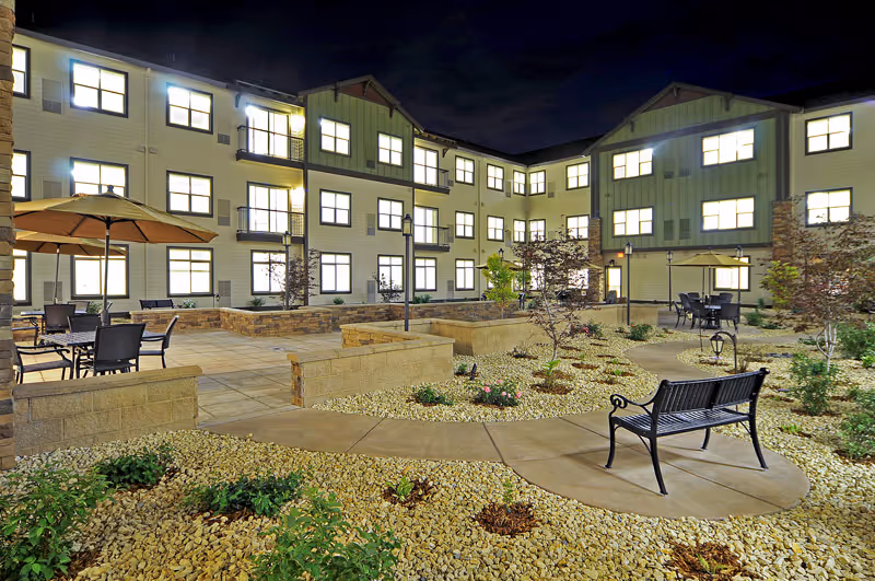Night view of an outdoor courtyard area in a senior living facility with illuminated windows of a three-story building surrounding the courtyard. The courtyard features paved walkways, small landscaped garden beds with rocks and plants, benches, tables with umbrellas, and chairs.