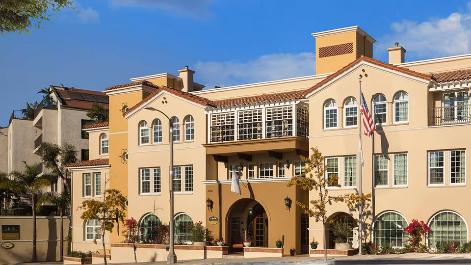 Exterior view of a multi-story senior living facility building with beige and tan walls, arched windows, a balcony, and an American flag on a flagpole in front. The building is surrounded by small trees and plants under a clear blue sky.
