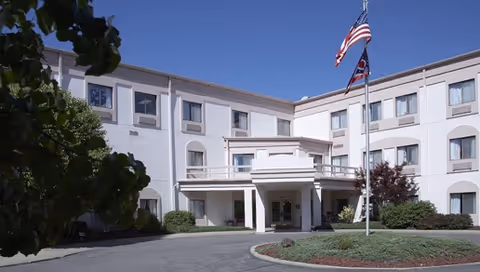 Exterior view of a three-story senior living facility building with a covered entrance, landscaped bushes, and two flagpoles displaying the American flag and another flag under a clear blue sky.