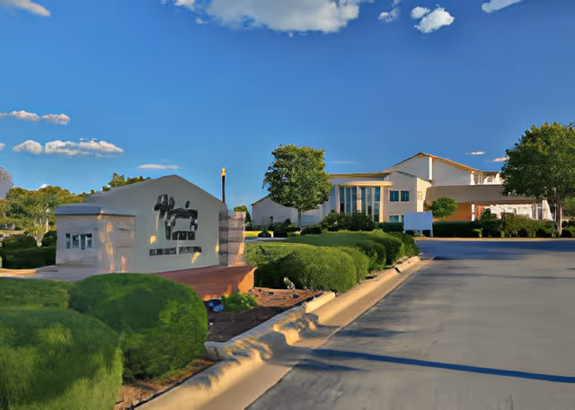 Exterior view of a senior living facility with a clear blue sky, manicured bushes, a paved driveway, and a building in the background with large windows and a light-colored facade.