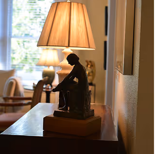 A small seated figurine sculpture on a wooden table with lamps and living-room furniture softly lit in the background.