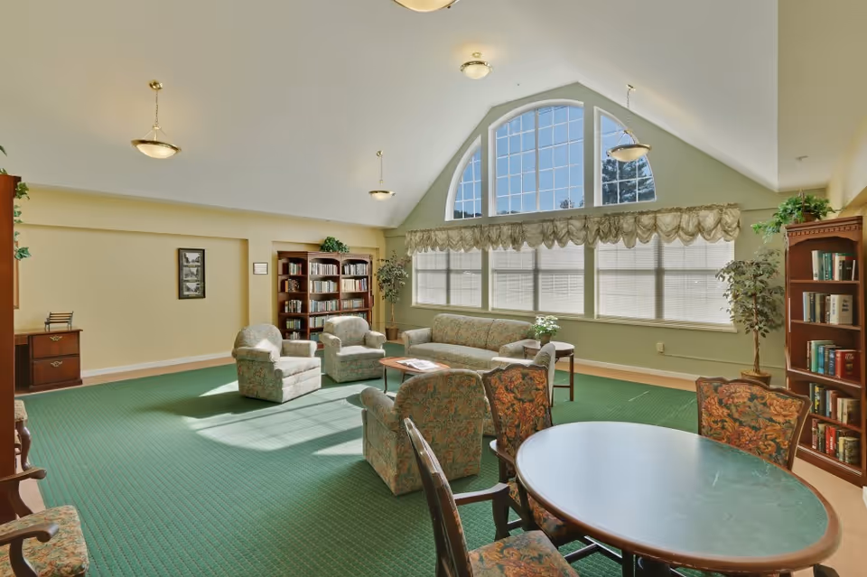 A spacious living room with green carpet, floral upholstered chairs and sofa, a round table with four chairs, bookshelves filled with books, large arched window with blinds and valance, and ceiling lights.