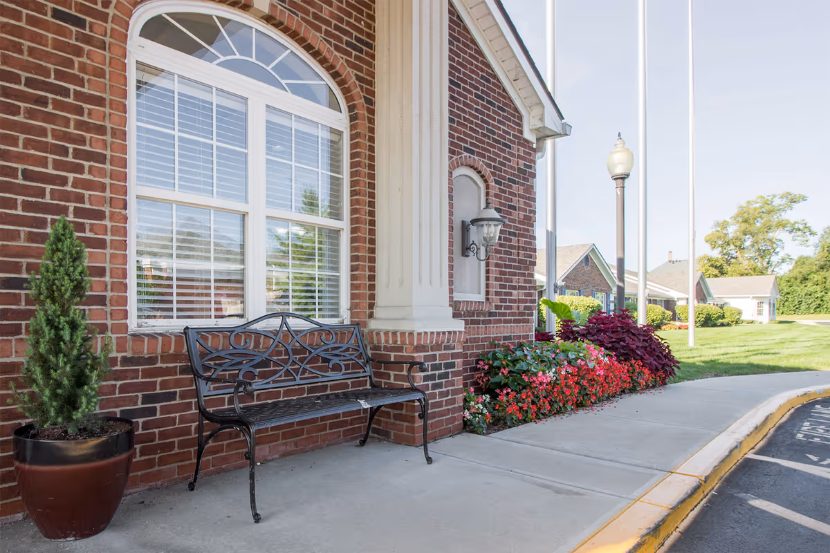 Brick building entrance with a decorative metal bench, potted evergreen, lamp, and colorful flowerbeds beside a sidewalk and flagpoles.