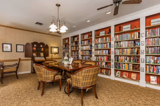 A cozy library room with a large wooden table surrounded by six plaid upholstered chairs. The back wall features built-in bookshelves filled with books, labeled with categories such as Fiction. The room has beige patterned carpet, warm beige walls, a ceiling fan, and a chandelier with five lights hanging above the table. There is also a wooden desk and chair in the corner with framed pictures on the wall.