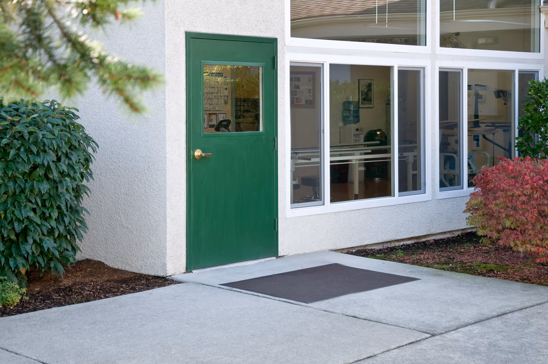A green exterior door and large windows on the side of a senior living facility with shrubs and a concrete walkway.