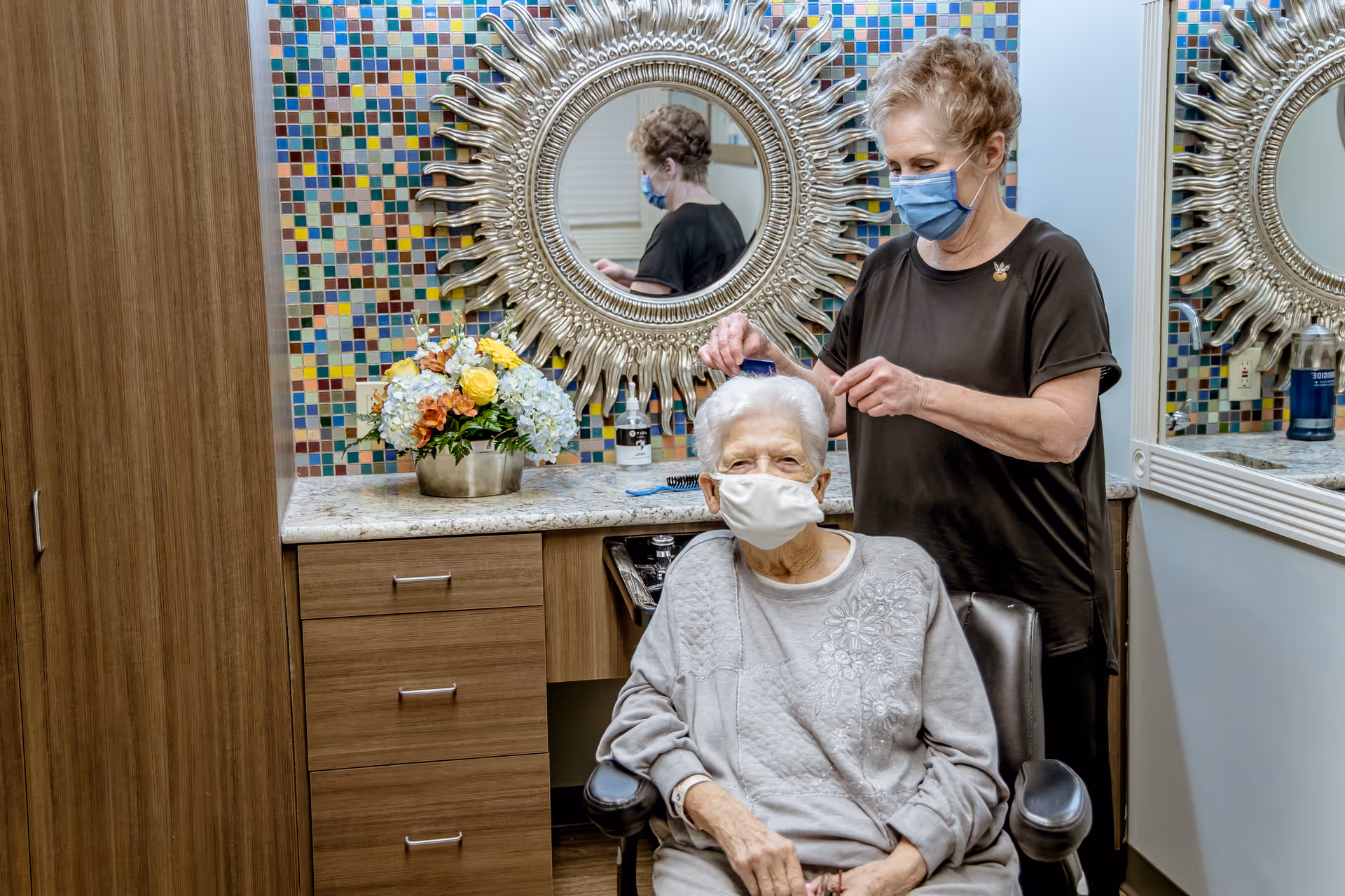 An elderly woman wearing a face mask is seated in a chair while another woman, also wearing a face mask, styles her hair in a room with a colorful tiled wall and a decorative round mirror. There is a countertop with a flower arrangement and hair care products.