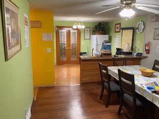 Interior view of a dining area with a wooden dining table and chairs. The table is covered with a patterned tablecloth and has various items on it. The room has green and yellow walls, wooden flooring, and ceiling fans with lights. In the background, there is a kitchen area with a refrigerator, cabinets, and a counter. A doorway leads to another room with wooden double doors and a chandelier.