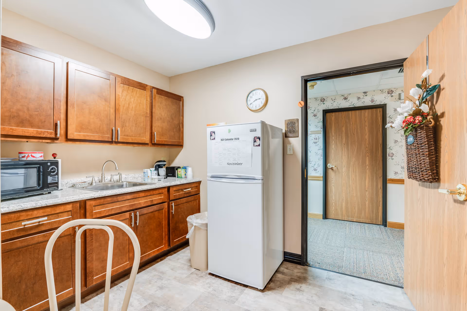 A small kitchen area with wooden cabinets, a countertop with a sink, a microwave, and various small appliances. There is a white refrigerator with a calendar on the door, a wall clock above it, and a trash can beside it. The door to the kitchen is open, showing a hallway with floral wallpaper and a closed wooden door.