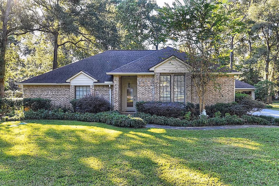 Single-story brick house with a front lawn, shrubs, and trees in a wooded setting.
