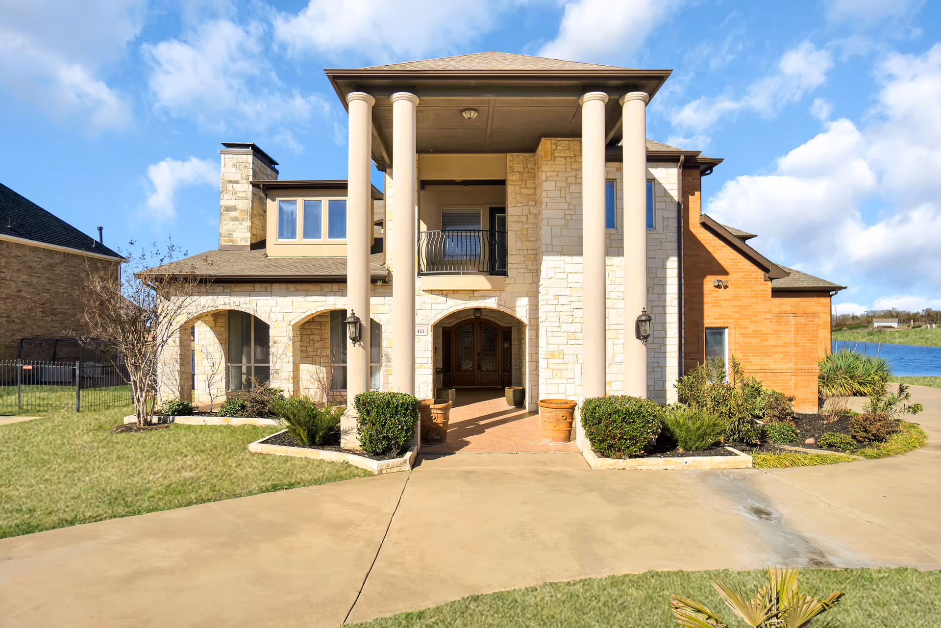 Front exterior view of a two-story residential building with large columns, stone and brick facade, a balcony above the entrance, and a driveway leading up to the front door. The sky is clear with some clouds.