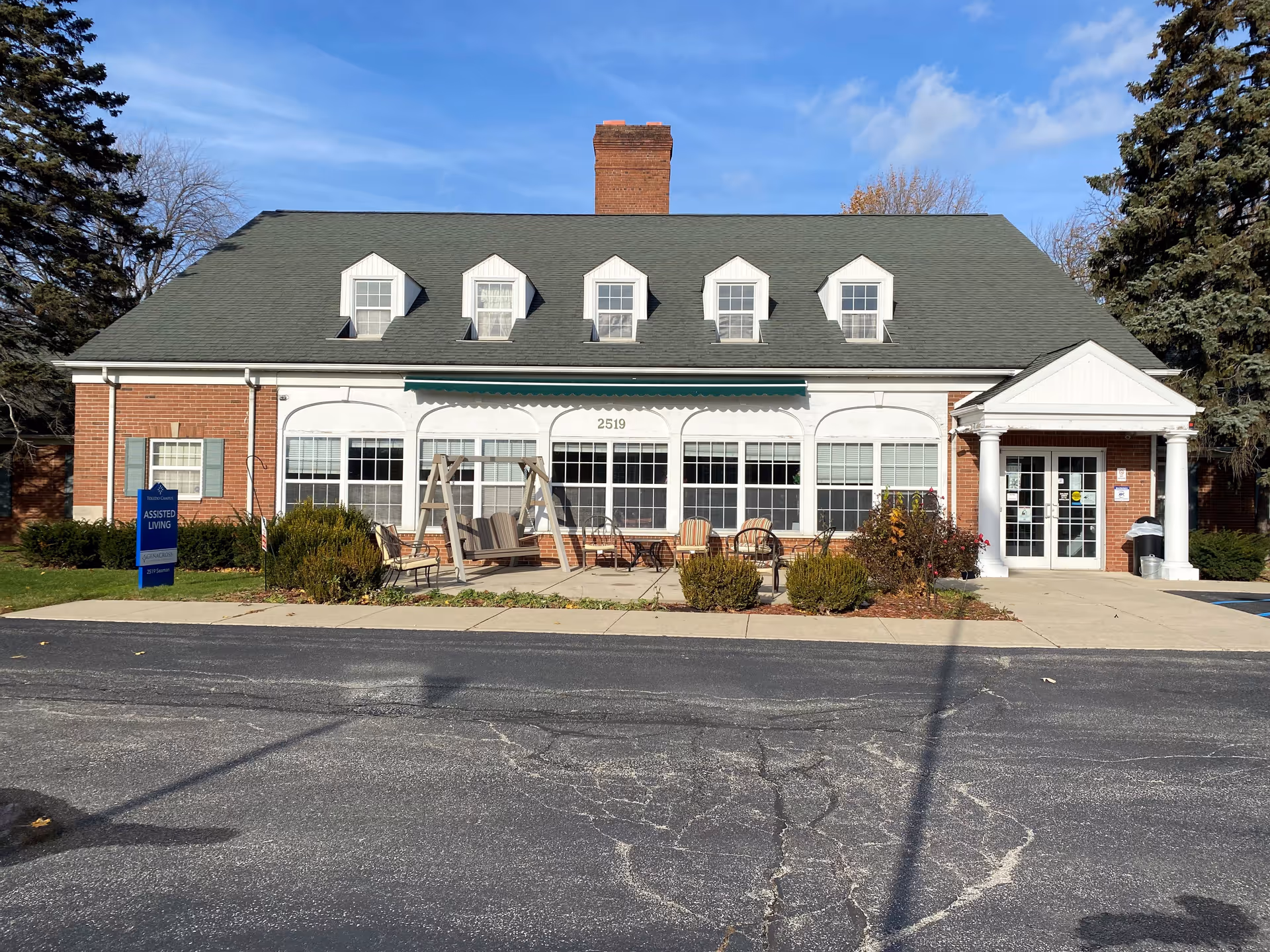 Front exterior view of a brick assisted living facility building with a green roof, five dormer windows, a central chimney, and a white porch entrance supported by columns. There is a patio area with outdoor seating and a swing in front of large windows. A blue sign on the left side reads 'Toledo County Assisted Living'.